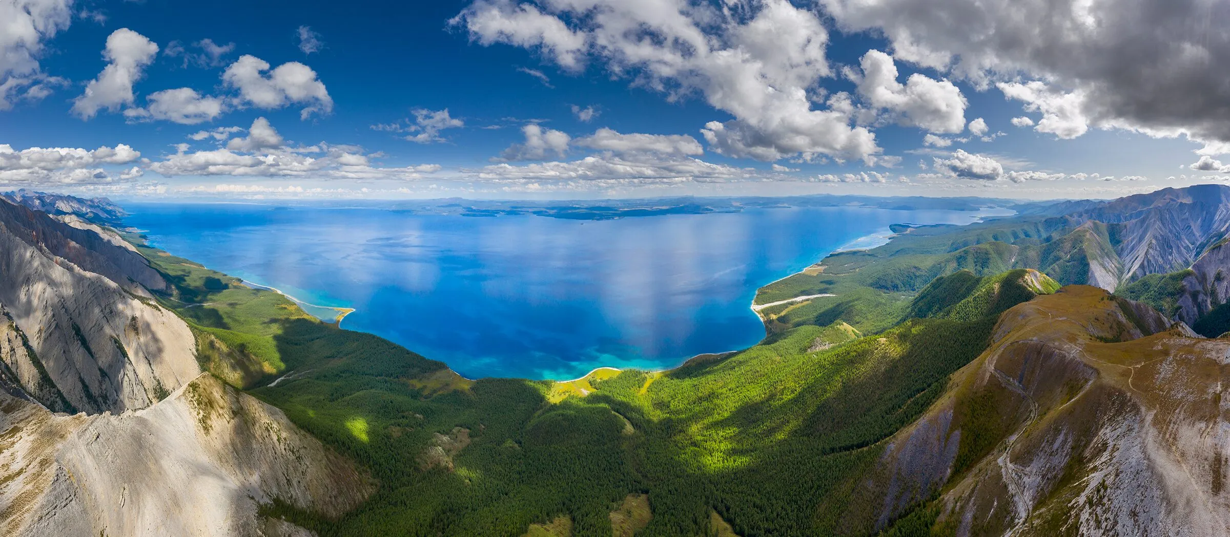 Aerial panorama of a deep blue lake set among forested mountains in northern Mongolia, cumulus clouds drifting overhead.
