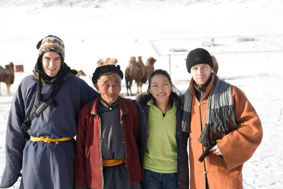 Baska in green, standing in the snow with a Mongolian elder and two foreign guests, Bactrian camels behind.
